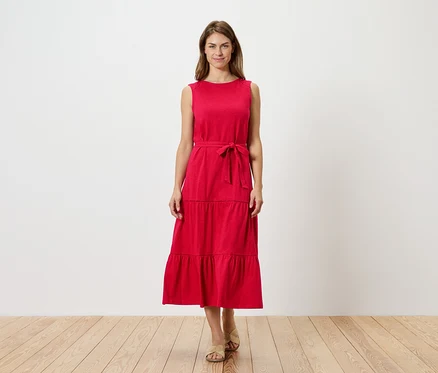 Une femme se tient devant un mur blanc et porte une robe rouge sans manches à plusieurs niveaux avec une ceinture et des sandales.