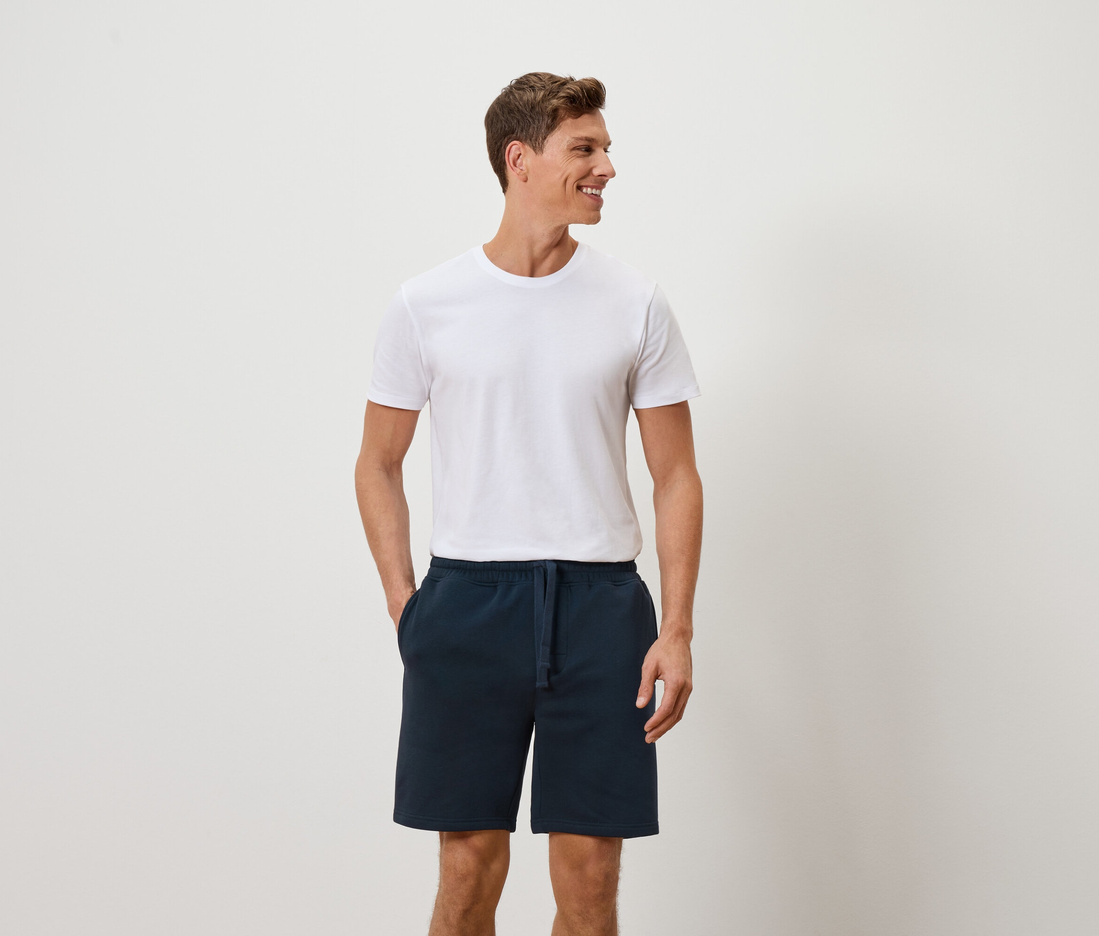 Homme en t-shirt blanc et short bleu, les mains dans les poches, souriant sur le côté.