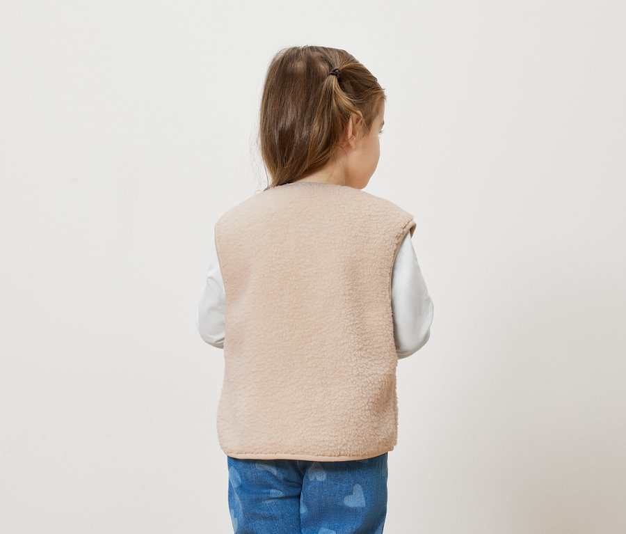 Vue arrière d'une petite fille avec une queue de cheval, portant un gilet en polaire beige sur une chemise à manches longues blanche et un jean bleu avec des motifs de cœurs.