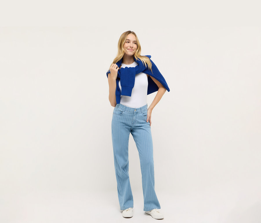 Femme posant en t-shirt blanc et jean bleu Angels Denim « Liz » Wide Leg, avec un pull bleu sur les épaules.