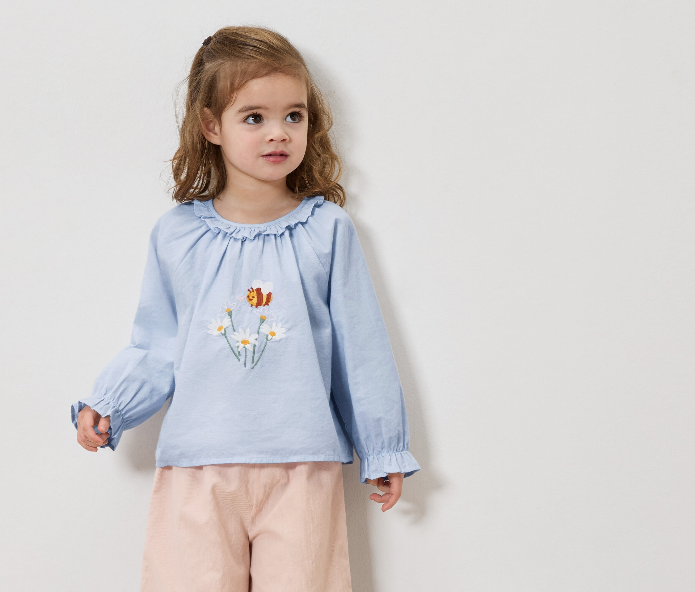 Une fillette aux cheveux blonds porte un chemisier bleu clair avec des broderies de marguerites et d'abeilles et un pantalon rose.