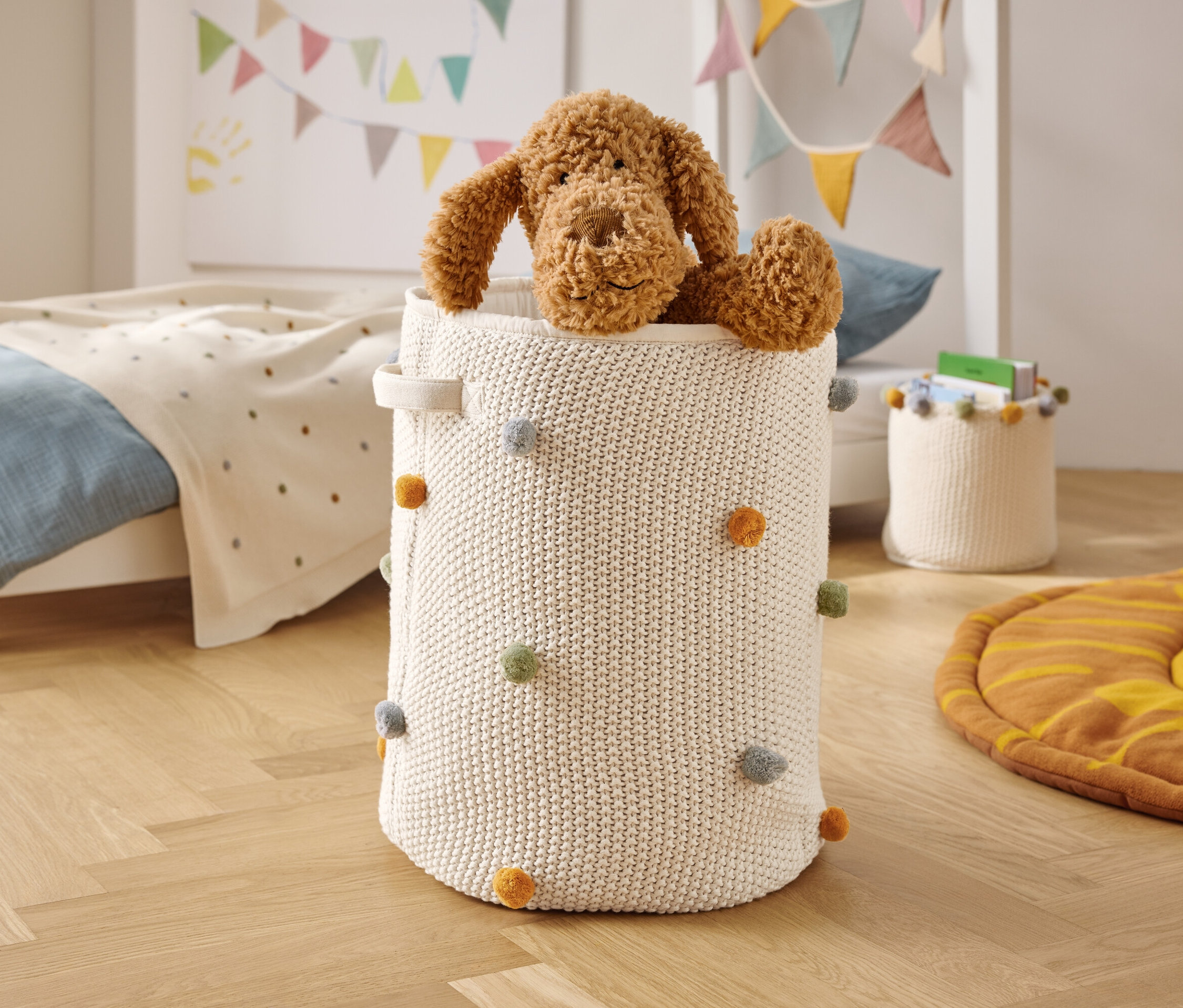 Un chien en peluche marron est assis dans un panier tricoté blanc avec des pompons colorés.