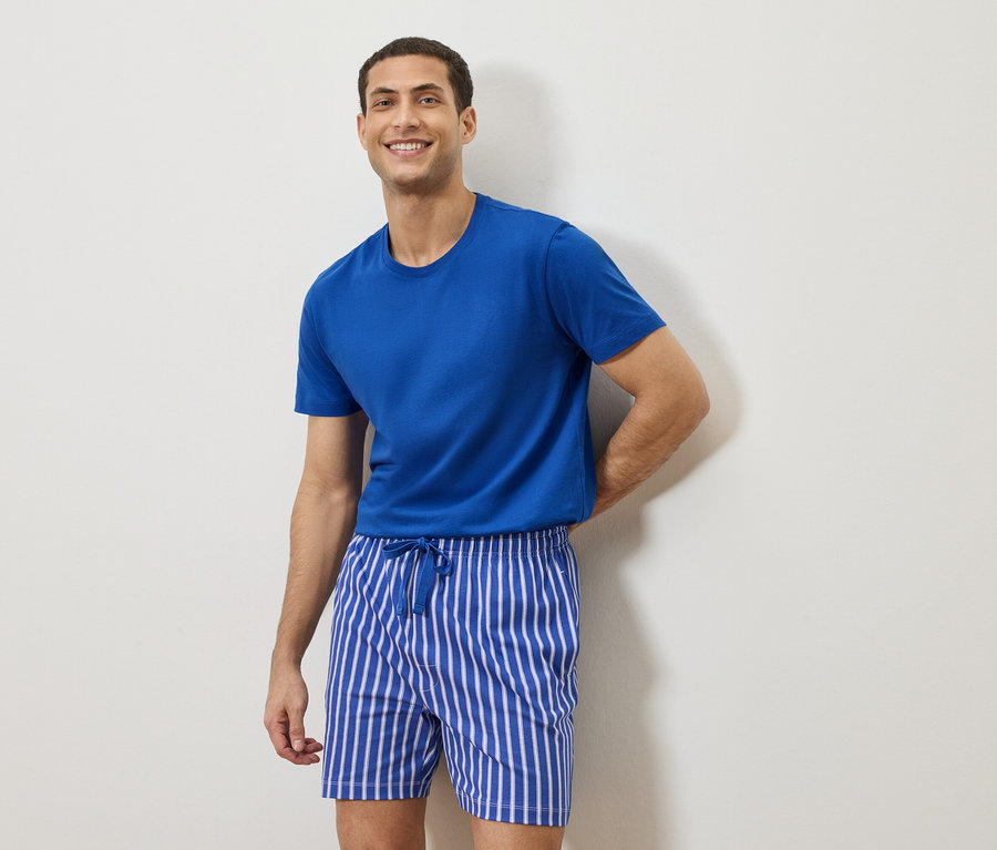 Homme portant un t-shirt bleu et un short rayé debout devant un mur blanc.