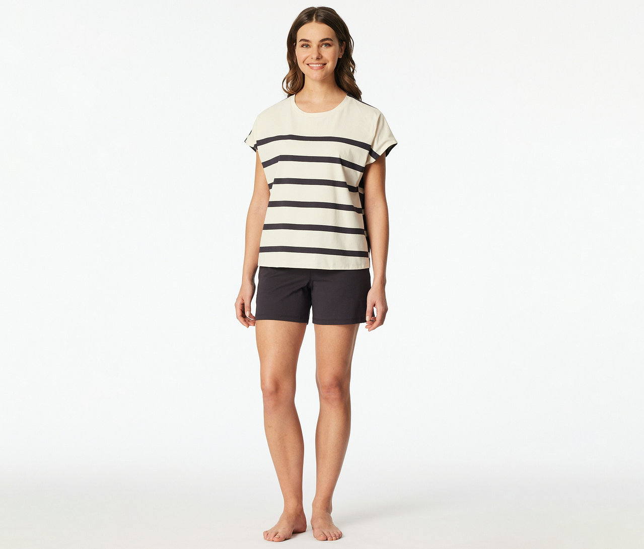 Femme souriante portant un set shorty SCHIESSER: un haut à manches courtes rayé crème et un short gris foncé. Elle est pieds nus.