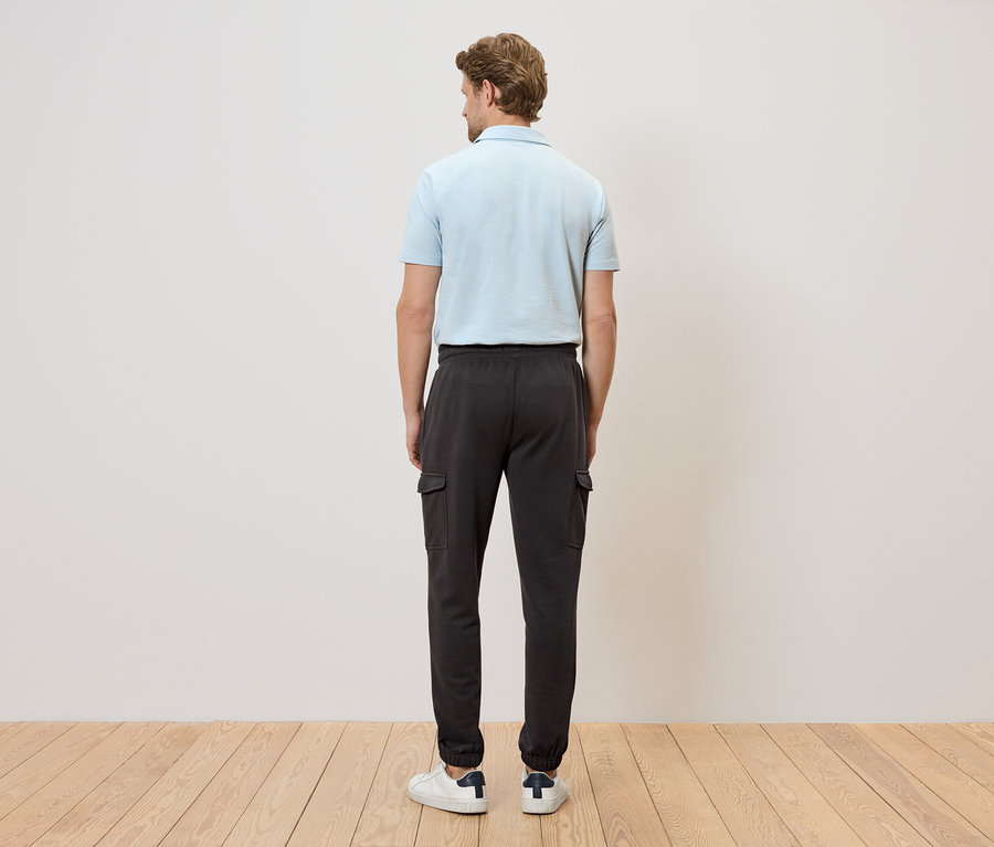Homme vu de dos portant un polo bleu clair et un pantalon cargo foncé devant un mur blanc et un plancher en bois.