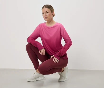 Une femme accroupie porte des vêtements de sport : un haut rose et rouge à manches longues, un legging bordeaux et des baskets gris clair sur fond blanc.