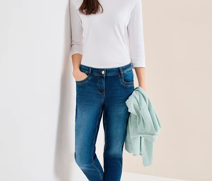 Femme portant un haut blanc à manches longues, une casquette de baseball bleue et un jean CECIL « Scarlett », appuyée contre un mur, tenant une chemise rayée vert clair.