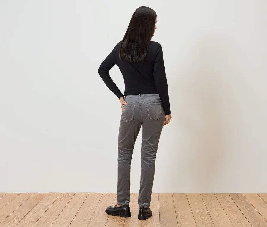 Une femme aux longs cheveux foncés se tient dos à la caméra, portant un haut noir à manches longues, un pantalon en velours côtelé gris et des chaussures noires.