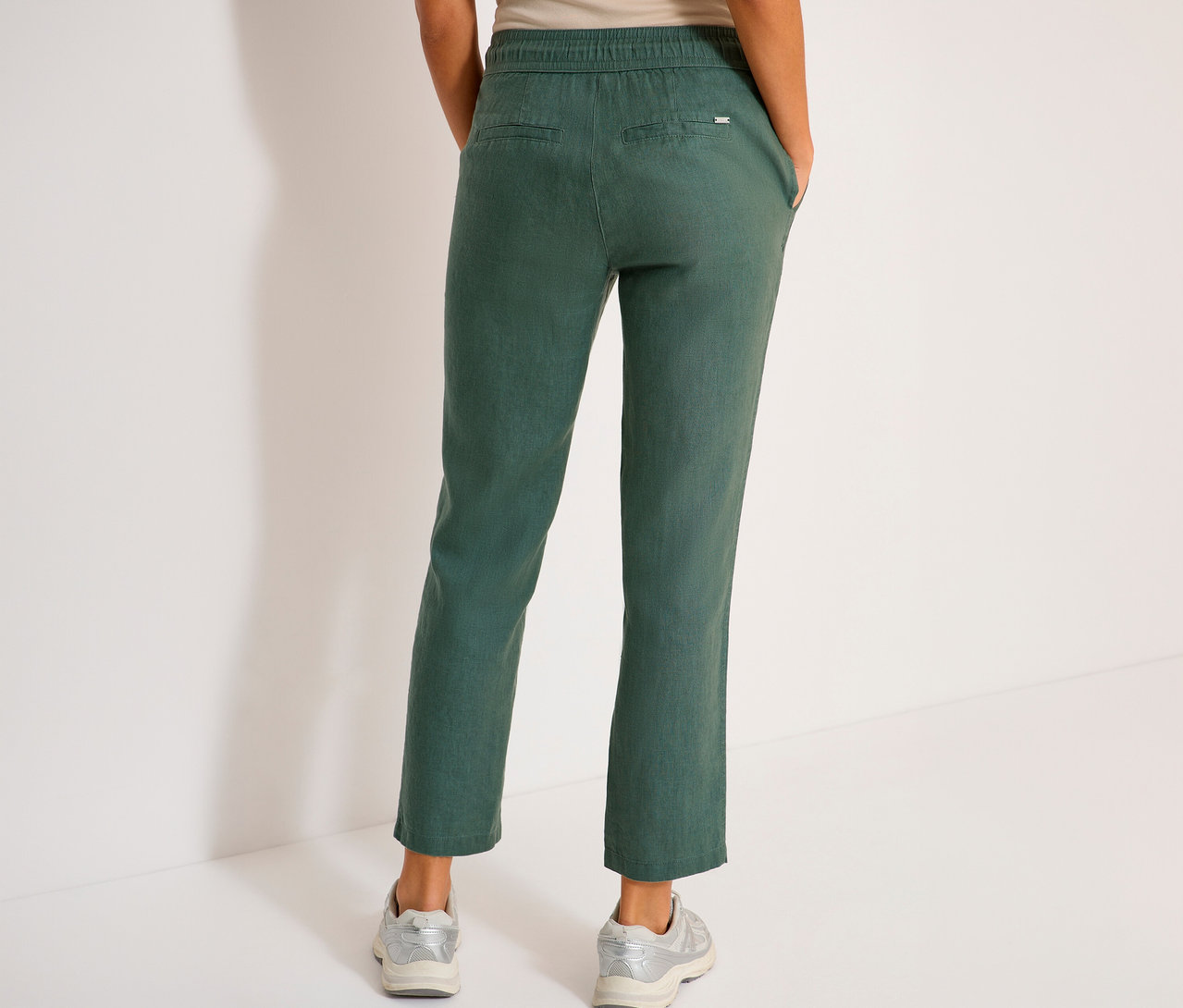 Vue arrière d'une femme portant un pantalon vert et des baskets argentées sur fond blanc.