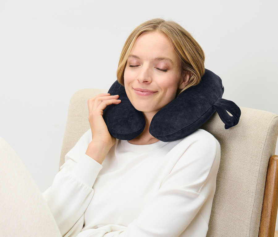 Une femme est assise sur une chaise, les yeux fermés, et porte un coussin de voyage bleu foncé.