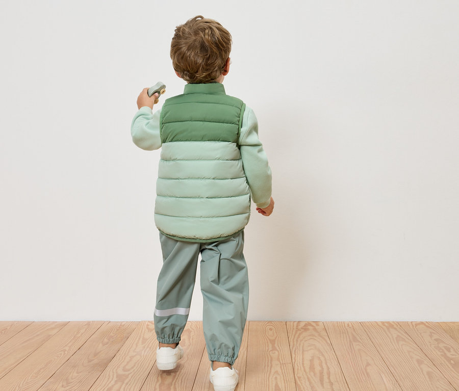 Un enfant vu de dos, portant un gilet matelassé vert clair et un pantalon vert clair, debout sur un plancher en bois devant un mur blanc.