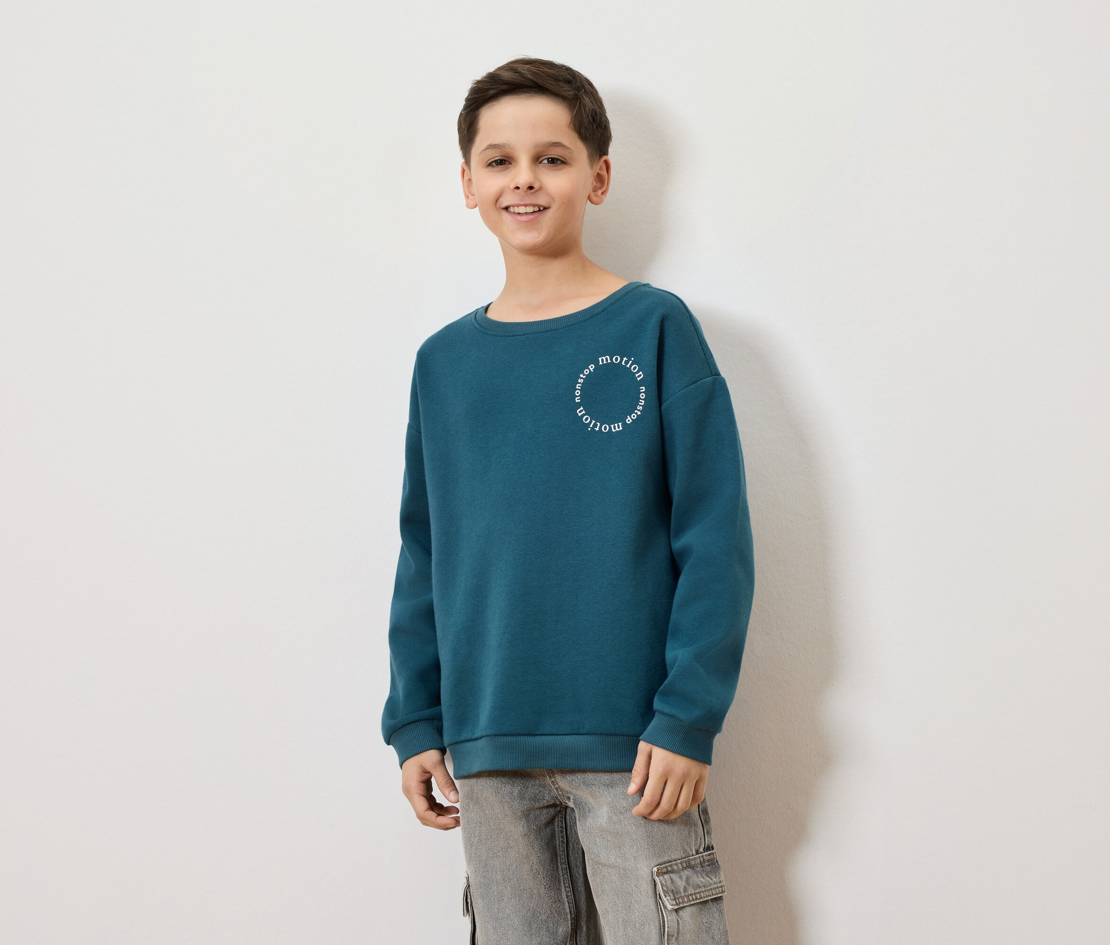 Un garçon souriant porte un sweat-shirt bleu-vert avec l'inscription "nonstop motion" et un pantalon cargo gris sur un fond blanc.