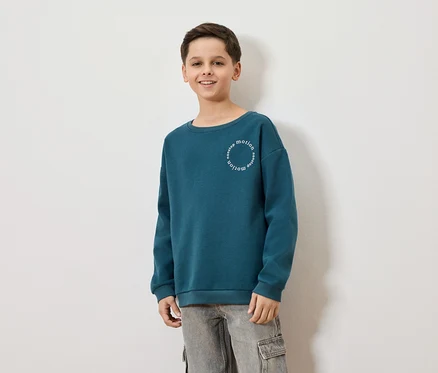 Un garçon souriant porte un sweat-shirt bleu-vert avec l'inscription "nonstop motion" et un pantalon cargo gris sur un fond blanc.