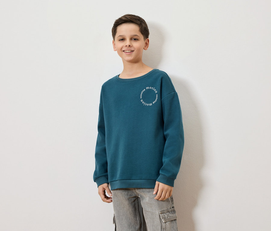 Un garçon souriant porte un sweat-shirt bleu-vert avec l'inscription "nonstop motion" et un pantalon cargo gris sur un fond blanc.