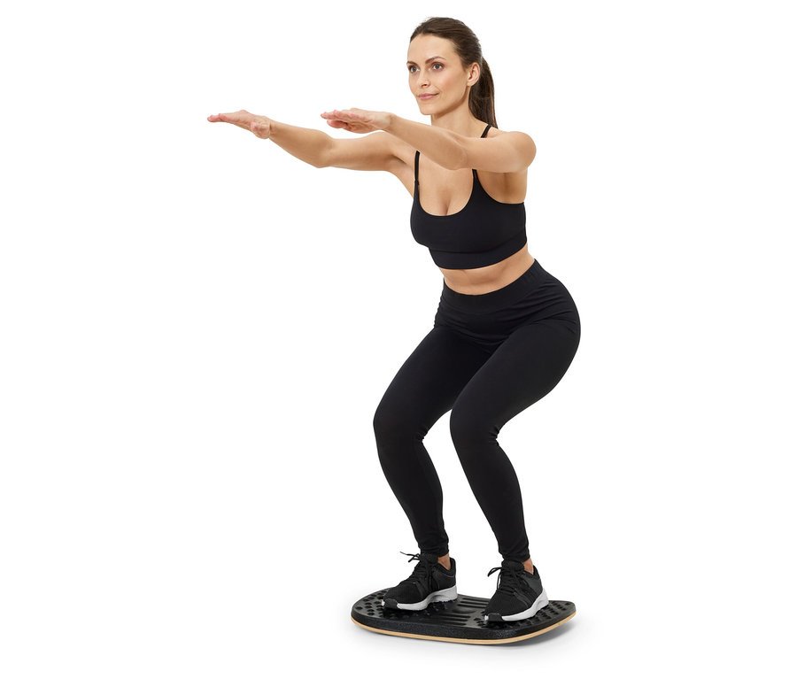 Femme en soutien-gorge de sport noir et leggings faisant des squats sur une planche d'équilibre.