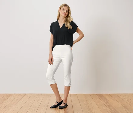 Une femme se tient debout, une main sur la hanche, portant un chemisier noir, un pantacourt blanc et des ballerines noires.