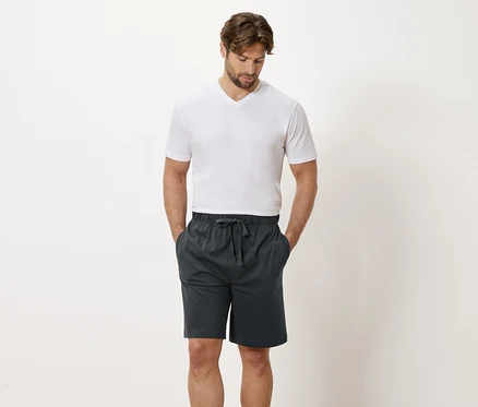 Un homme en t-shirt blanc et short gris se tient devant un mur blanc.