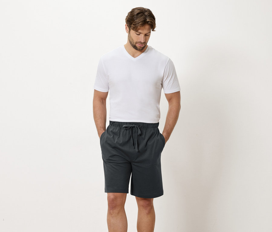 Un homme en t-shirt blanc et short gris se tient devant un mur blanc.