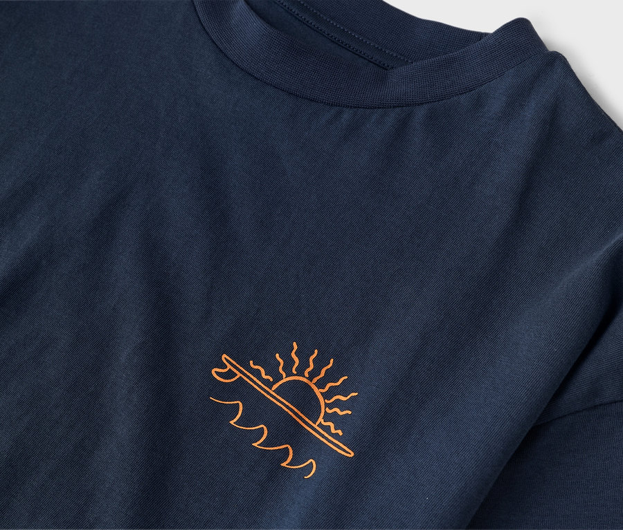 Gros plan sur un t-shirt bleu marine avec un imprimé orange représentant un soleil au-dessus d'une planche de surf et des vagues.