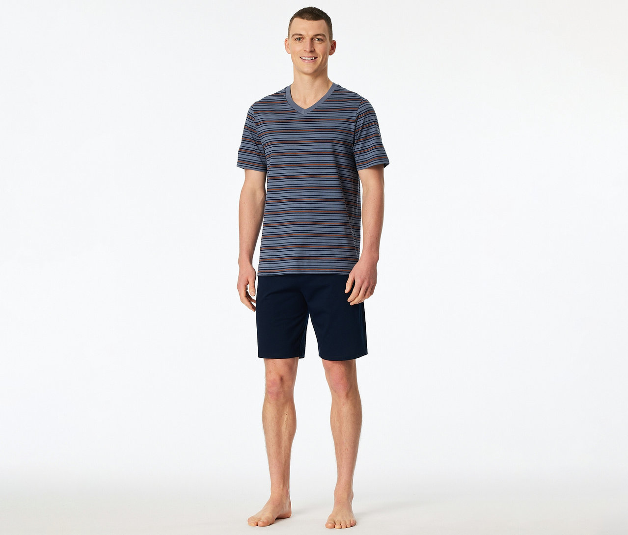 Un homme souriant se tient pieds nus, vêtu d'un set shorty SCHIESSER rayé à col en V et d'un short bleu marine.