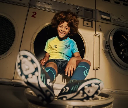 Un garçon souriant avec une coupe afro est assis dans une machine à laver, portant un maillot jaune et bleu avec le numéro 12, un short, des chaussettes et des chaussures de football.