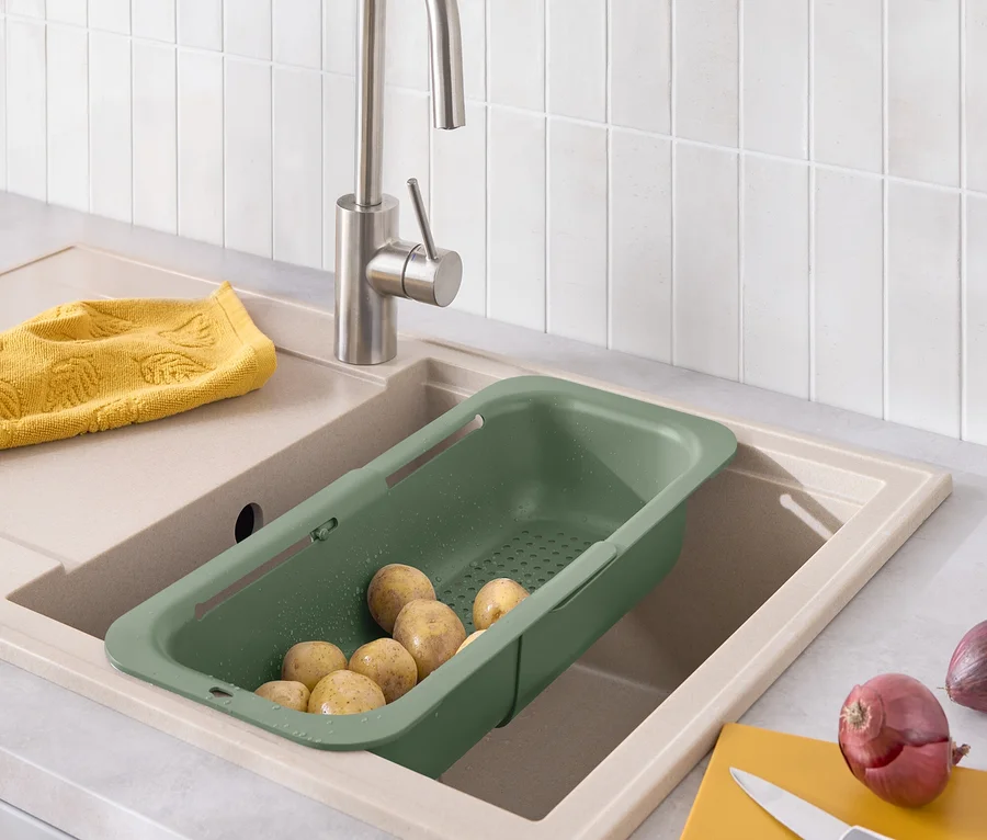 Des pommes de terre dans une passoire verte placée dans un évier, à côté d'un torchon jaune, d'une planche à découper jaune et d'un oignon.