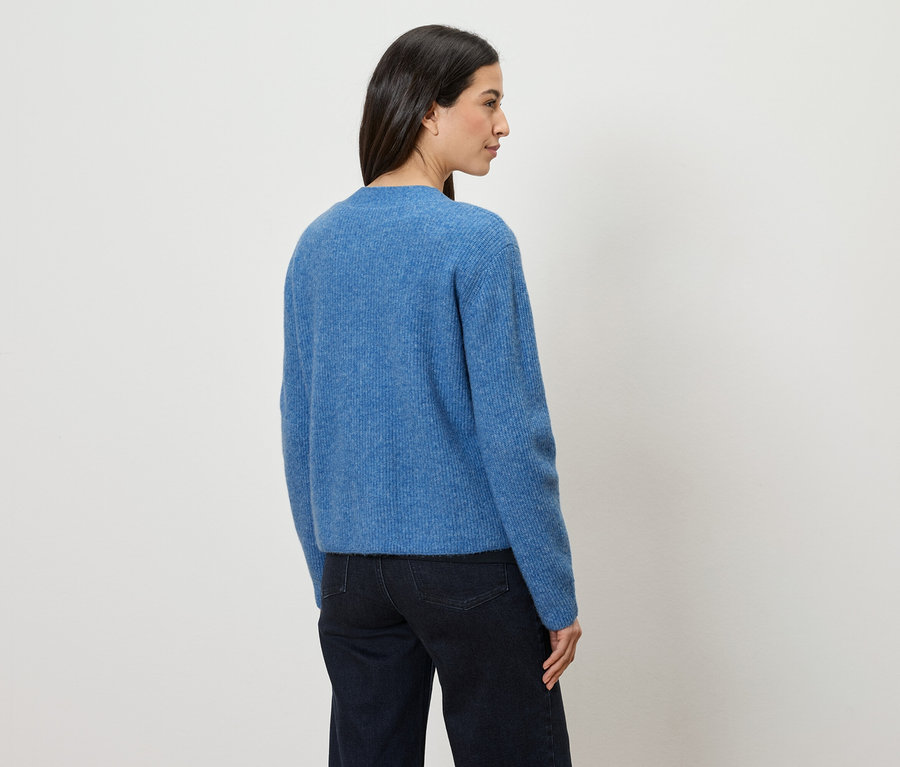 Une femme aux cheveux foncés porte un pull bleu et un jean foncé, se tenant devant un mur blanc.