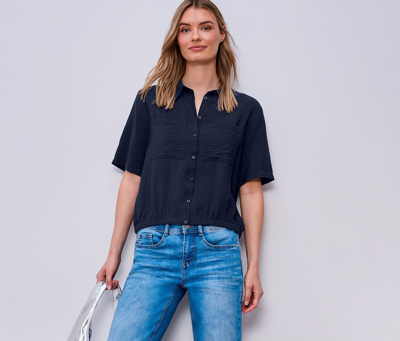 Une femme porte un jean bleu et un chemisier Street One. Elle tient un sac argenté.