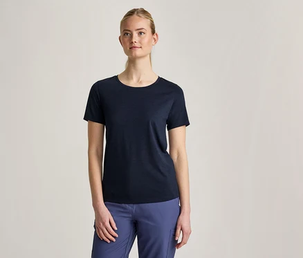 Une femme aux cheveux blonds attachés porte un t-shirt bleu foncé à manches courtes et un pantalon bleu, debout devant un fond gris clair.