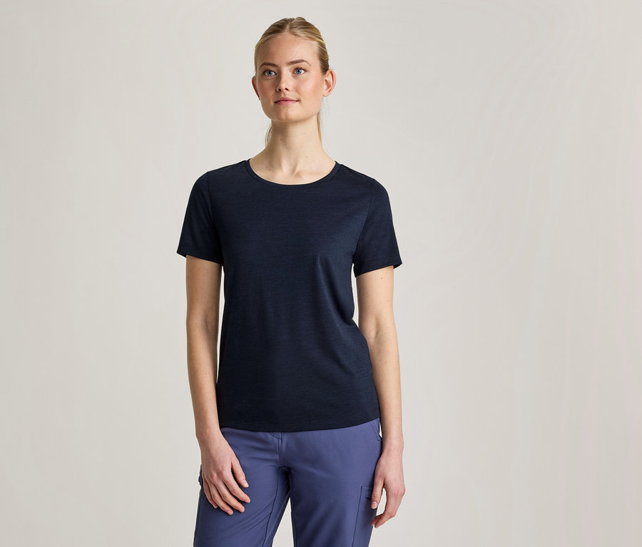 Une femme aux cheveux blonds attachés porte un t-shirt bleu foncé à manches courtes et un pantalon bleu, debout devant un fond gris clair.