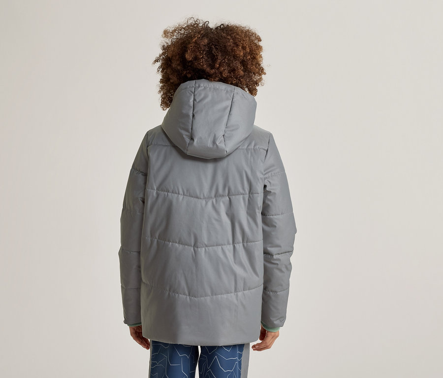 Personne aux cheveux bouclés vue de dos, portant une veste grise matelassée à capuche et un pantalon bleu à motifs.