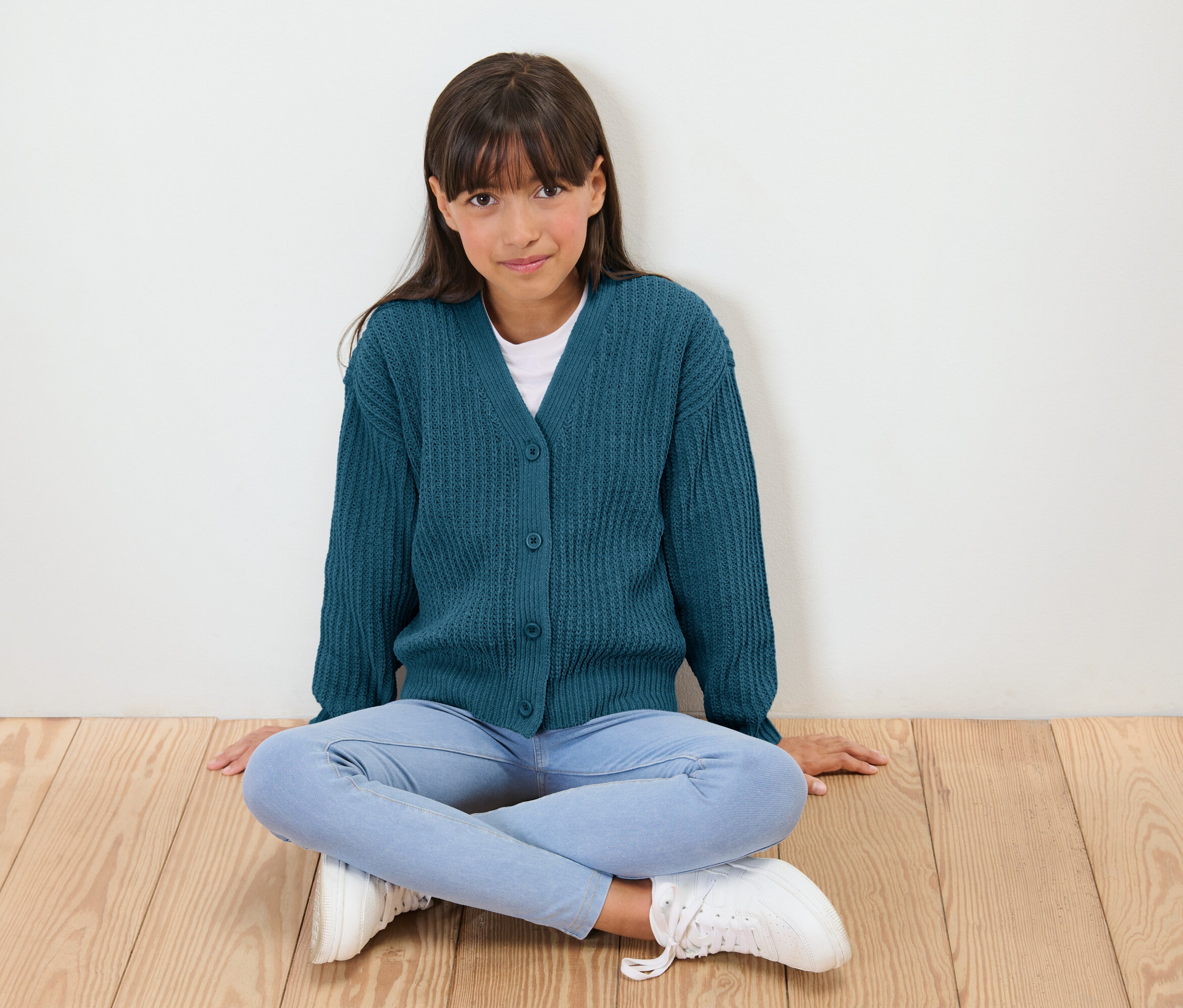 Une jeune fille est assise les jambes croisées sur un plancher en bois, portant un cardigan bleu, un chemisier blanc, un jean et des baskets blanches.