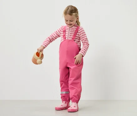 Une fille verse de l'eau avec un arrosoir, portant une salopette rose et des bottes.