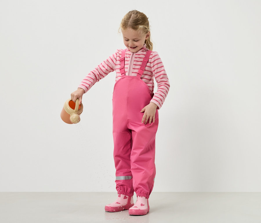 Une fille verse de l'eau avec un arrosoir, portant une salopette rose et des bottes.