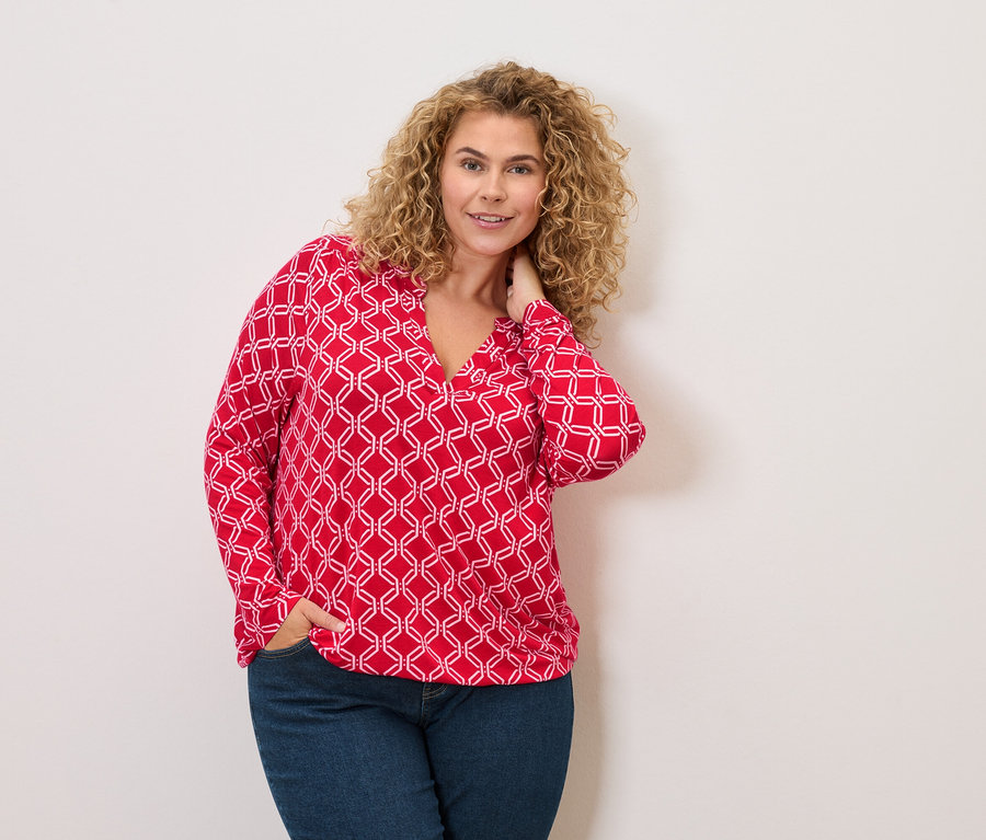 Femme aux cheveux bouclés portant un chemisier rouge et blanc et un jean, posant avec une main dans sa poche.