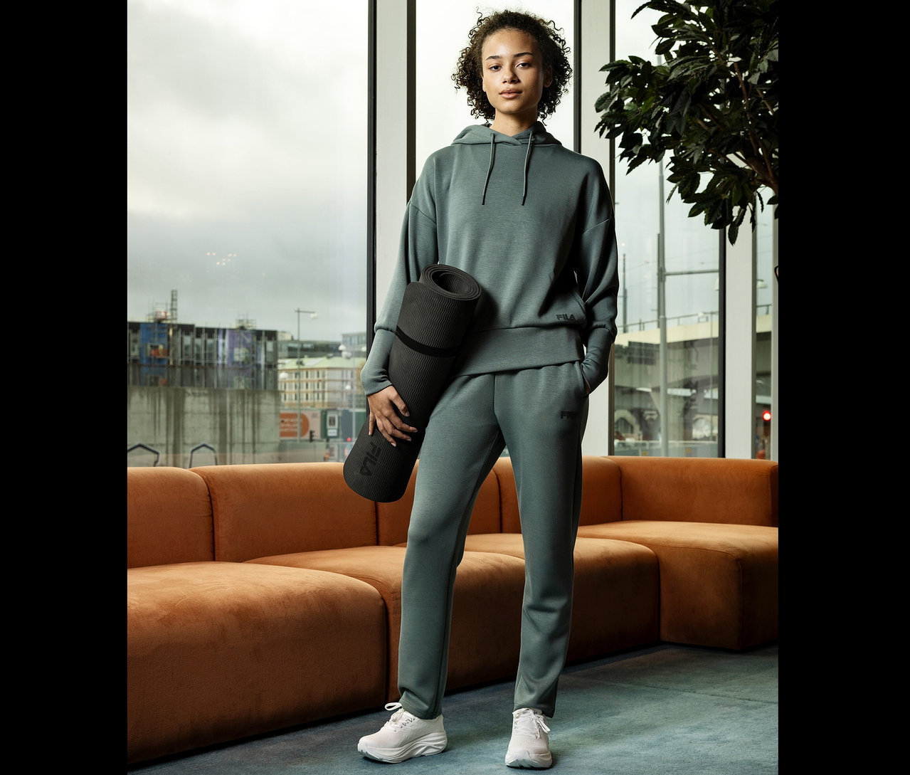 Jeune femme en survêtement gris tenant un tapis de yoga enroulé devant une fenêtre.