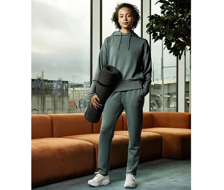 Femme portant un hoodie pour femme FILA en mélange de modal couleur sauge et un pantalon de jogging assorti, tenant un tapis de yoga noir, debout dans une pièce moderne avec de grandes fenêtres.