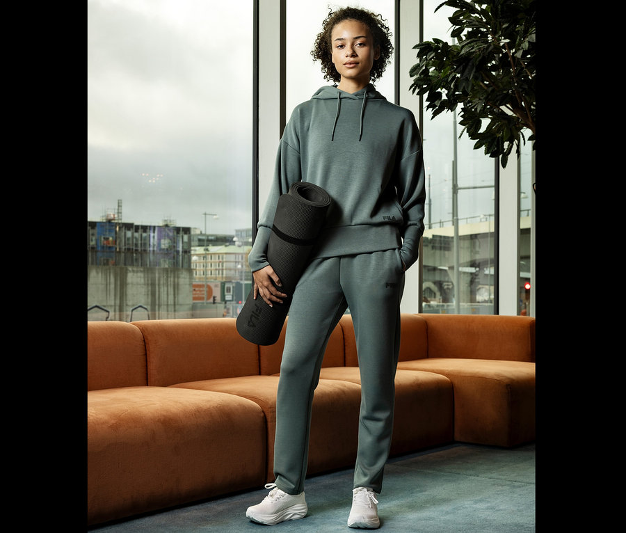 Jeune femme en survêtement gris tenant un tapis de yoga enroulé devant une fenêtre.