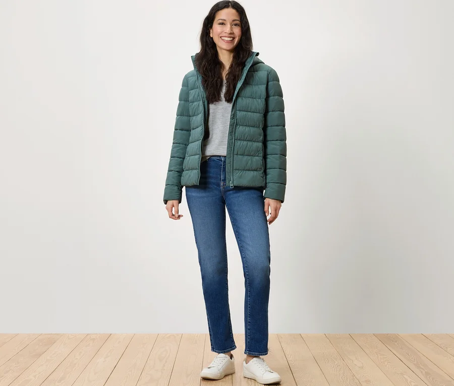 Une femme se tient debout et sourit, portant une doudoune verte, un jean bleu et des baskets blanches.