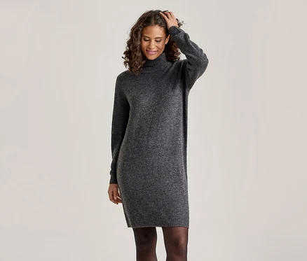 Une femme souriante aux cheveux bouclés porte une robe pull à col roulé gris foncé et des collants noirs, une main touchant ses cheveux.