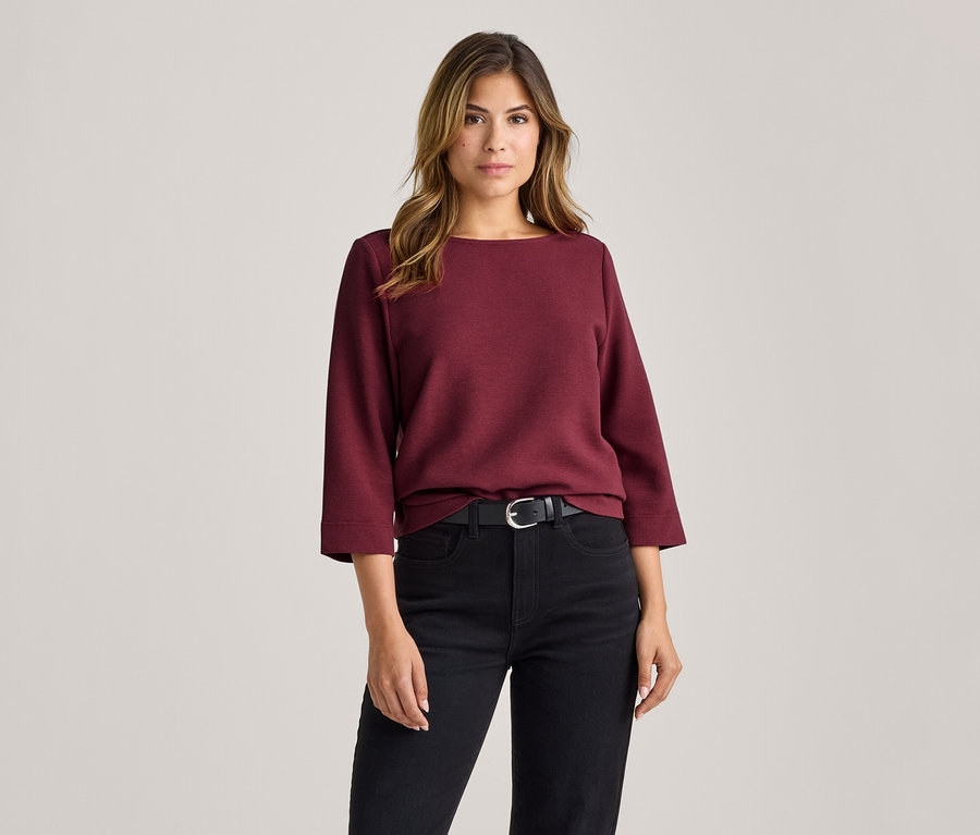Femme portant un haut bordeaux à col bateau avec des manches 3/4, un jean noir et une ceinture noire à boucle argentée.