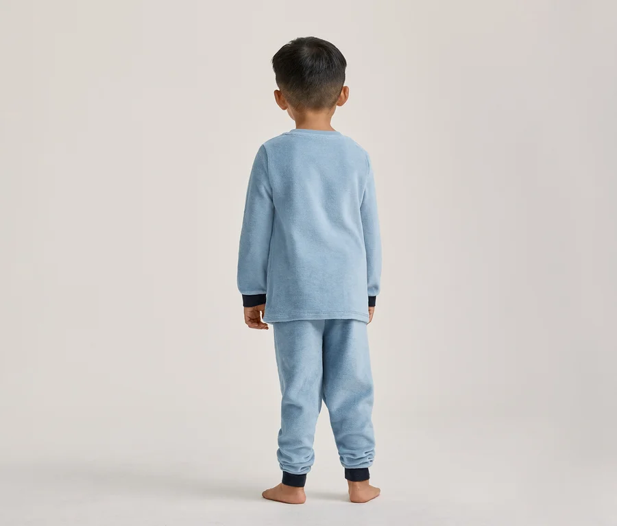 Enfant de dos portant un pyjama bleu clair avec des poignets et chevilles bleu foncé, debout pieds nus.