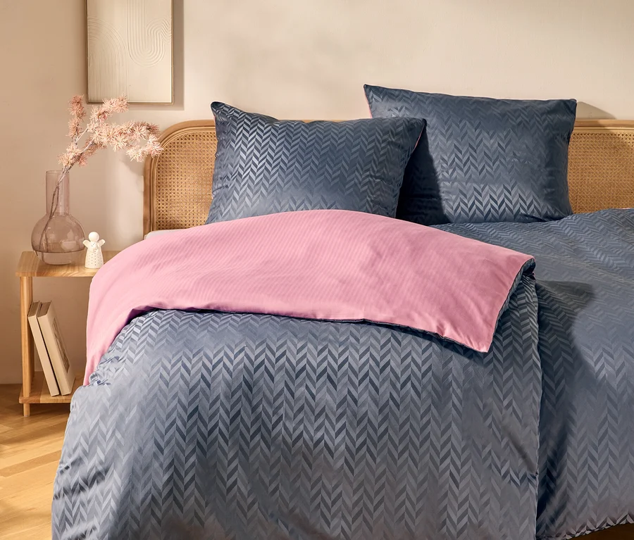 Lit avec tête de lit en rotin, parure de lit bleu foncé à motif chevrons et jeté de lit rose. À côté, une table de chevet en bois avec un vase, des fleurs roses et des livres. Une œuvre d'art encadrée est accrochée au mur.