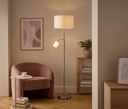 Un coin salon confortable avec un lampadaire argenté doté d'une lumière principale et d'une liseuse, un fauteuil rembourré rose et une table d'appoint avec une tasse. Des œuvres d'art murales et une étagère en bois avec une plante sont également visibles.
