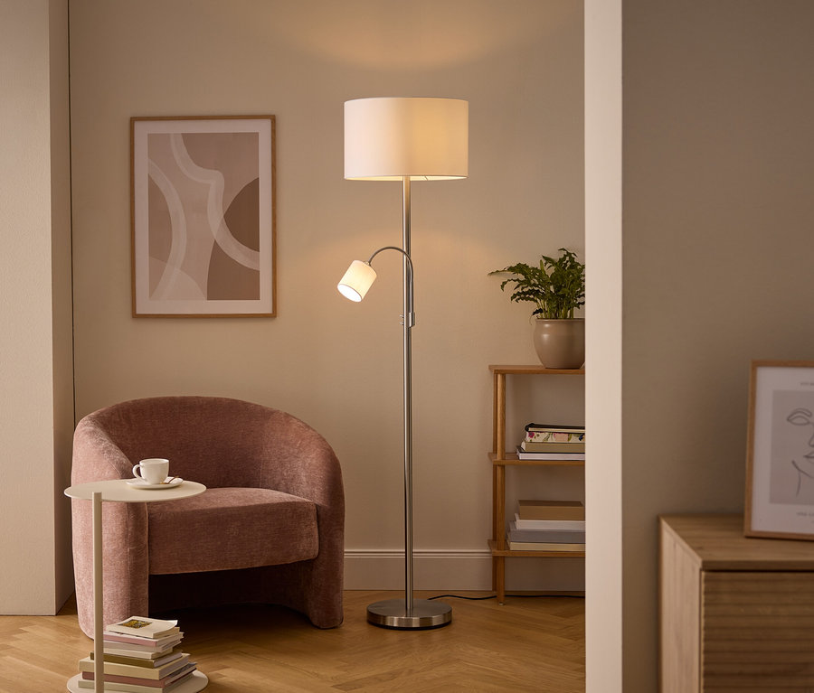 Un coin salon confortable avec un lampadaire argenté doté d'une lumière principale et d'une liseuse, un fauteuil rembourré rose et une table d'appoint avec une tasse. Des œuvres d'art murales et une étagère en bois avec une plante sont également visibles.