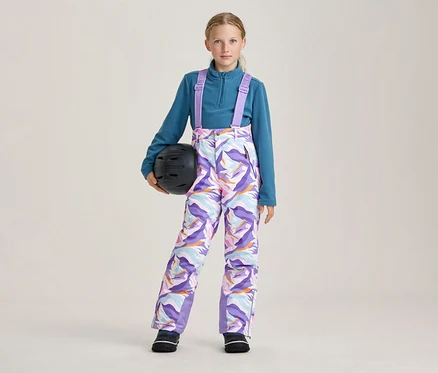 Jeune fille portant un haut en polaire sarcelle et un pantalon de ski à motifs colorés avec des bretelles violettes, tenant un casque de ski noir dans sa main gauche.