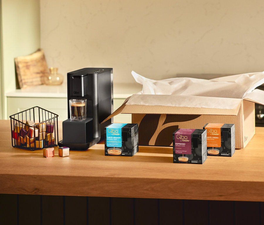 Machine à café noire Qbo Iconic, Dark Chrome avec un espresso dans un verre sur un comptoir en bois. À côté, un panier de capsules colorées, deux capsules détachées et trois boîtes, dont Qbo Caffè Grande KIRINYAGA BAYA, devant un carton ouvert.