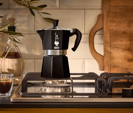 Une cafetière à expresso noire est posée sur une cuisinière à gaz à côté d'un verre d'expresso.