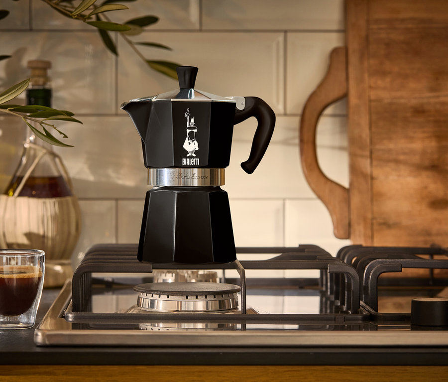 Une cafetière à expresso noire est posée sur une cuisinière à gaz à côté d'un verre d'expresso.