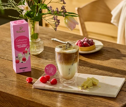 Sur une table en bois, un verre de café, des morceaux de chocolat blanc et des framboises sont posés sur une planche de marbre. À l'arrière-plan, on peut voir un paquet de Cafissimo Flavoured Espresso – White Choc & Raspberry – 10 capsules.